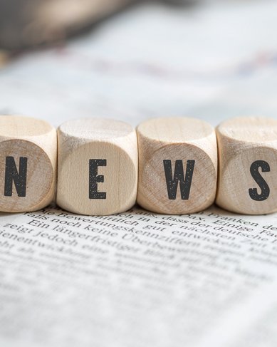 Holzklötze mit den Buchstaben "N", "E", "W", "S" auf einer Zeitungsoberfläche. Die Buchstaben sind deutlich sichtbar und stehen für Nachrichten. Der Hintergrund zeigt Teile einer Zeitung.