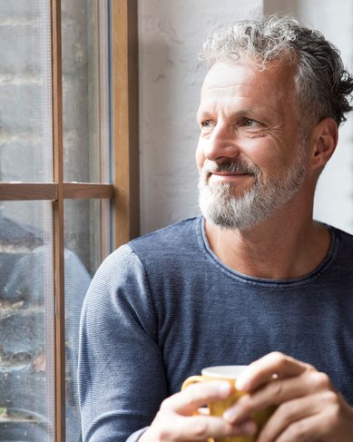 Ein Mann mit grauem Bart und lockigem Haar sitzt an einem Fenster und hält eine Tasse in der Hand. Er blickt nachdenklich nach draußen.