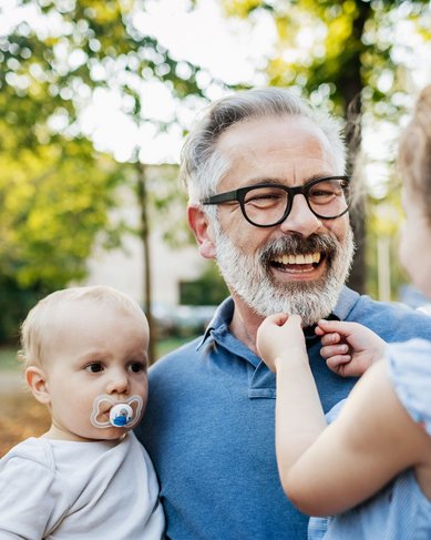 Ein älterer Mann mit Brille lächelt, während er ein Kleinkind auf dem Arm hält und ein weiteres Kind ihm gegenüber steht. Die Szene spielt sich in einem parkähnlichen Umfeld mit Bäumen ab.