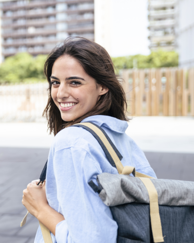Junge Frau mit einem Rucksack, die lächelnd über die Schulter schaut. Sie trägt ein hellblaues Hemd und steht auf einem Gehweg in einer urbanen Umgebung mit modernen Gebäuden im Hintergrund.