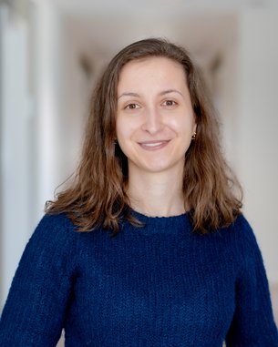 Eine lächelnde Frau mit langen, lockigen Haaren trägt einen blauen Pullover und steht in einem hellen Flur.