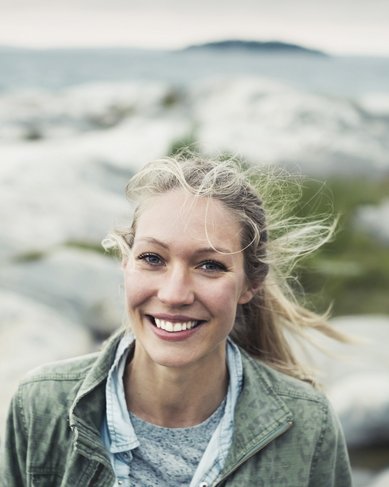 Eine lächelnde Frau mit langen, blonden Haaren steht auf Felsen am Meer. Im Hintergrund sind sanfte Hügel und eine bewölkte Himmel zu sehen.