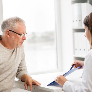 Ein älterer Mann mit Brille sitzt an einem Tisch und schaut aufmerksam auf eine Frau in einem weißen Kittel. Die Frau hält ein Dokument in der Hand und scheint Informationen zu präsentieren.