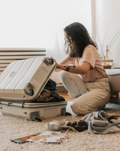 Eine Person sitzt auf dem Boden und packt einen Koffer mit Kleidung. Um sie herum liegen verschiedene Kleidungsstücke und Accessoires. Der Raum ist hell und modern eingerichtet.