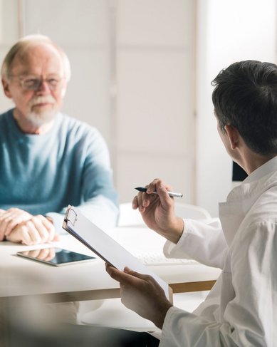 Ein älterer Mann sitzt an einem Tisch und spricht mit einem jüngeren Mann, der Notizen auf einem Clipboard macht. Auf dem Tisch steht ein Glas Wasser und ein Tablet.