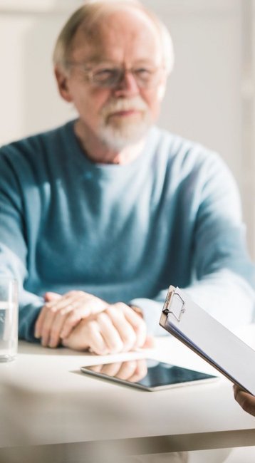 Ein älterer Mann sitzt an einem Tisch und spricht mit einem jüngeren Mann, der Notizen auf einem Clipboard macht. Auf dem Tisch steht ein Glas Wasser und ein Tablet.