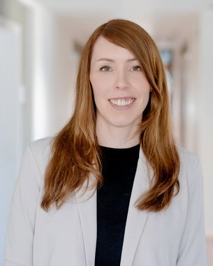 Eine lächelnde Frau mit langen, roten Haaren trägt einen hellen Blazer und steht in einem hellen Flur.