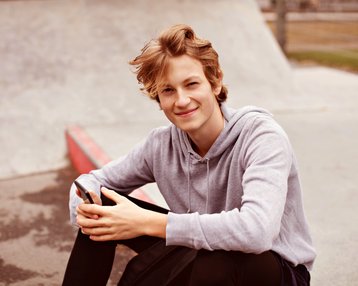 Ein junger Mann sitzt auf dem Boden eines Skateparks, lächelt und hält ein Smartphone in der Hand. Er trägt einen grauen Hoodie und hat lockiges, blondes Haar.