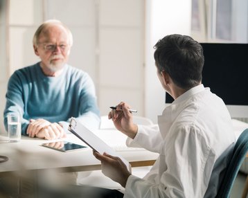 Suchtklinik: Ein älterer Mann sitzt an einem Tisch in Rehaklinik für Abhängigkeitserkrankungen und spricht mit einem jüngeren Mann, der Notizen auf einem Clipboard macht. Auf dem Tisch steht ein Glas Wasser und ein Tablet.